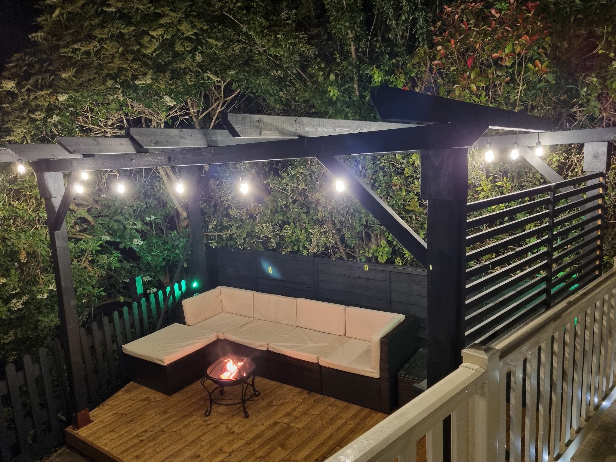 Garden pergola at night lit by festoon lights, with outdoor seating and a fire pit